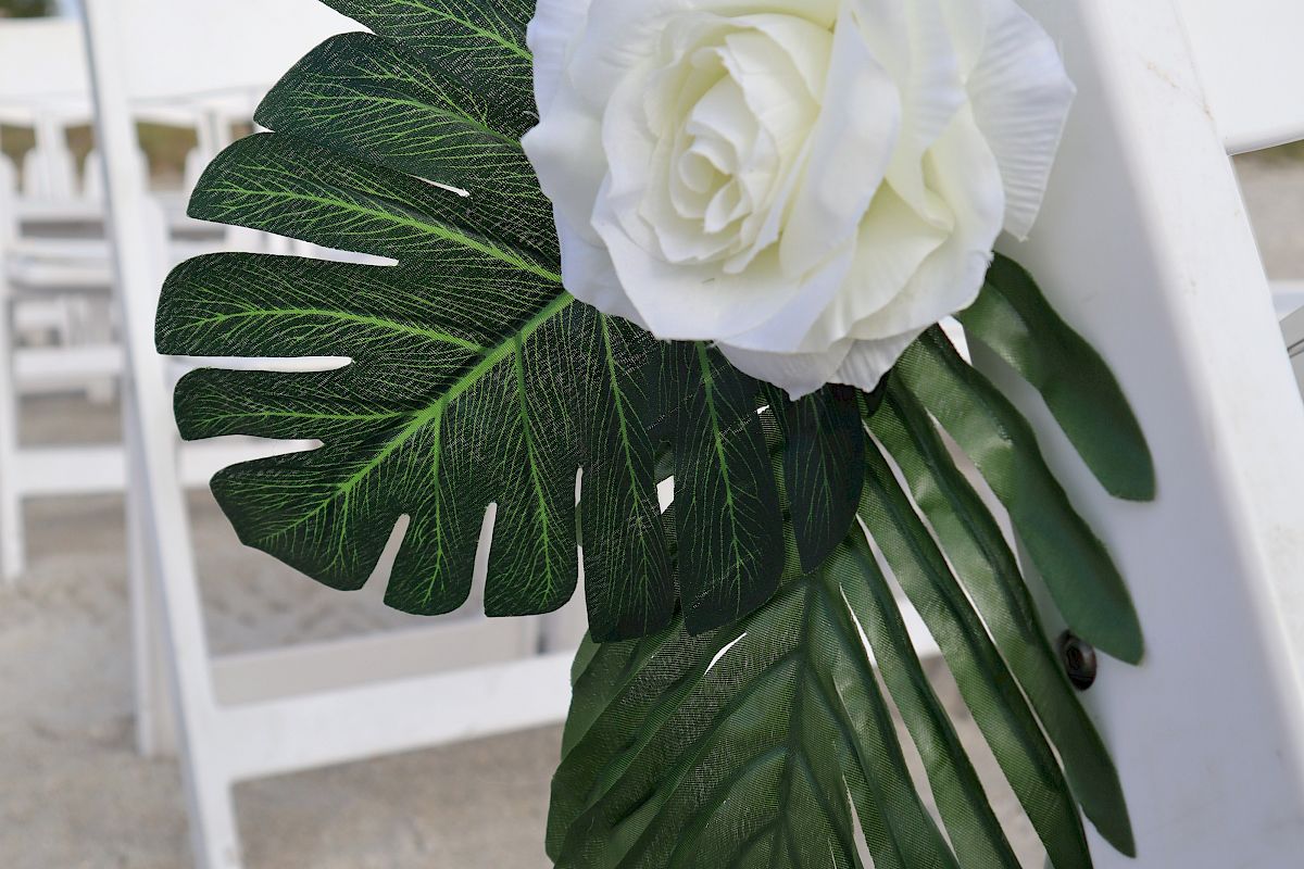 A white rose and green leaves are attached to a white wooden chair, set up outdoors, possibly for an event like a wedding, on a sandy surface.