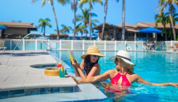 Two people relax in a pool with hats, near drinks on the edge, under a clear blue sky with palm trees in the background.