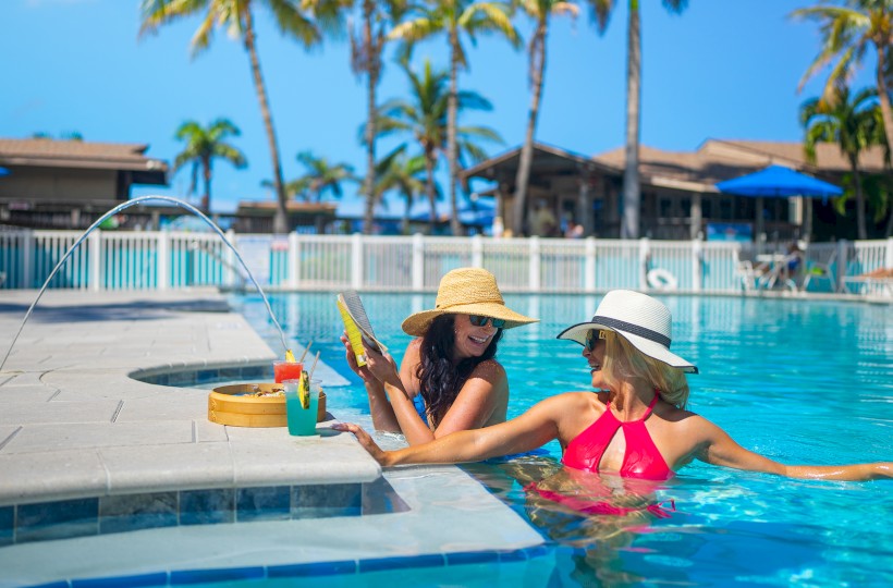 Two people relax in a pool with hats, near drinks on the edge, under a clear blue sky with palm trees in the background.