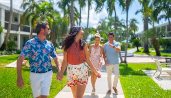 A group of four people walking outdoors, smiling and chatting, surrounded by palm trees and greenery, with a resort-like backdrop.