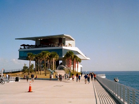 A pier with a structure at the end, visitors walking, clear skies, and calm sea.