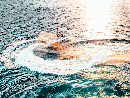 A person is riding a jet ski, creating a circular wave on a sunlit sea.