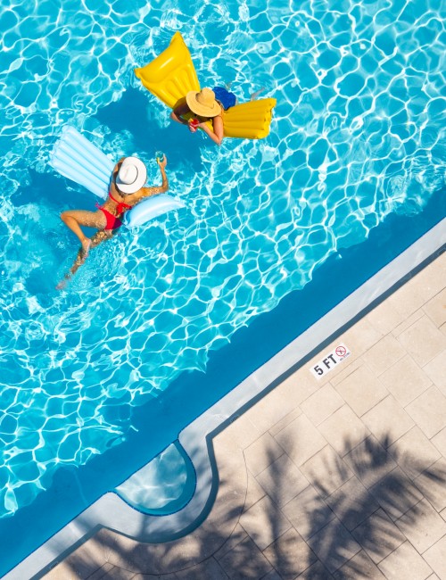 Two people relax on pool floats in a sunlit swimming pool, with a palm tree shadow on the deck.