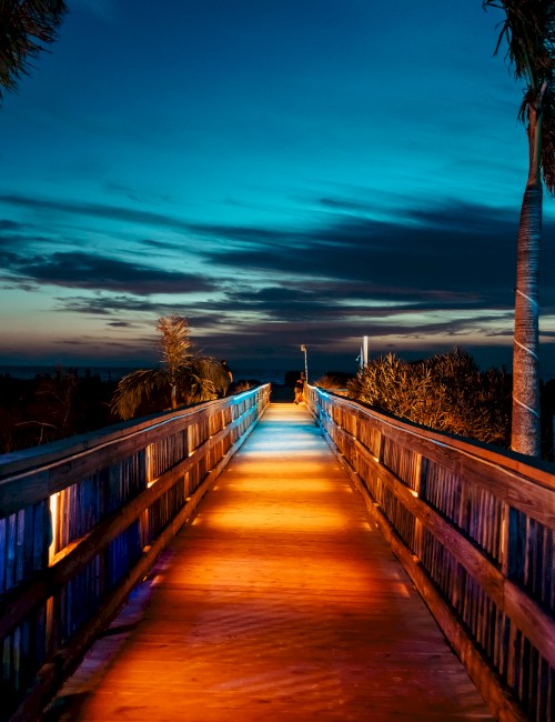 A wooden boardwalk illuminated by lights leads towards the horizon, with palm trees and a vibrant blue and orange sky at dusk.