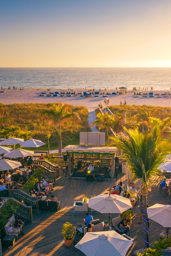 Beachfront scene with a bustling outdoor restaurant, white umbrellas, palm trees, and people enjoying the sunset by the ocean.