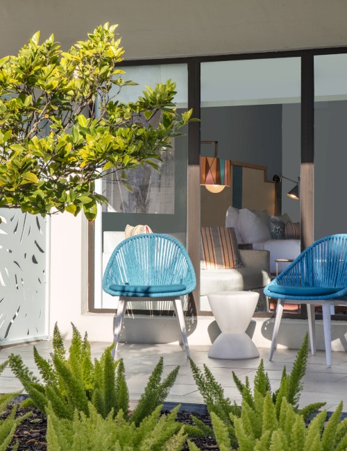 A patio with blue chairs and a small table is surrounded by greenery, with a view through large glass doors into a modern interior space.