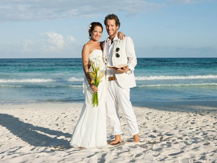 A couple in formal attire stands barefoot on a beach, with a clear blue ocean in the background, holding a bouquet.