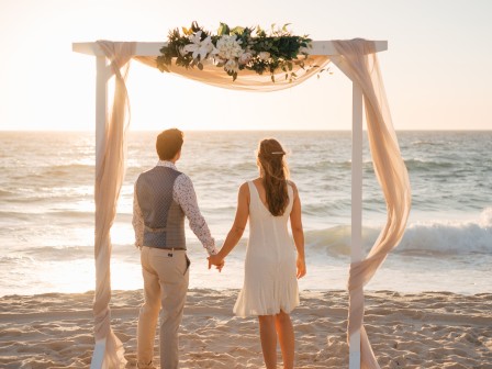 A couple stands under a decorated arch on a beach, holding hands and gazing at the ocean during sunset.