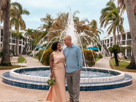 A couple stands in front of a fountain, surrounded by palm trees, with a romantic setting in the background.
