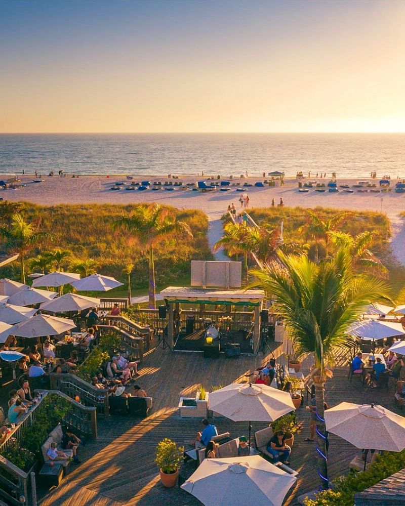 Beachside outdoor dining with umbrellas, people, and a view of the ocean during sunset.