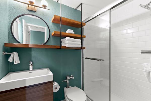 Modern bathroom with teal walls, mirror, sink, wooden shelves, toilet, and a glass shower enclosure.