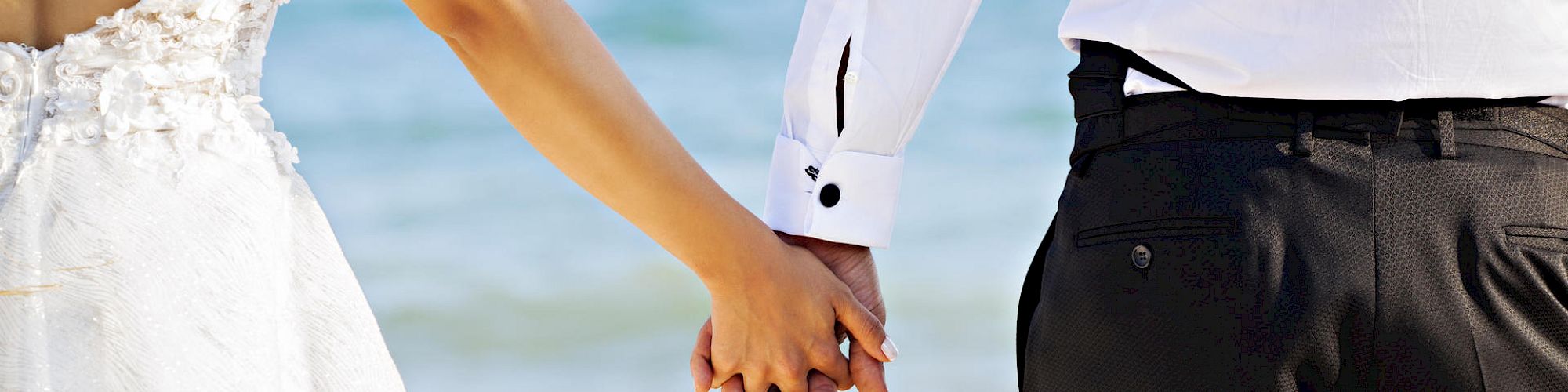 A couple is holding hands at the beach; the bride is in a wedding dress and the groom is in a dress shirt and pants.