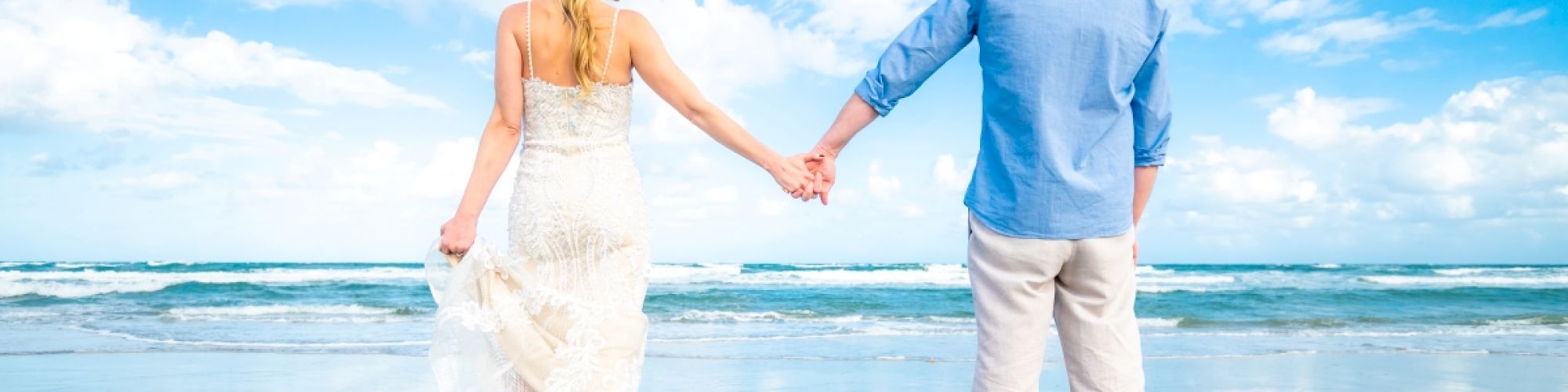 A couple holds hands while standing on a sandy beach, facing the ocean, with reflections on the wet sand and a bright blue sky above.