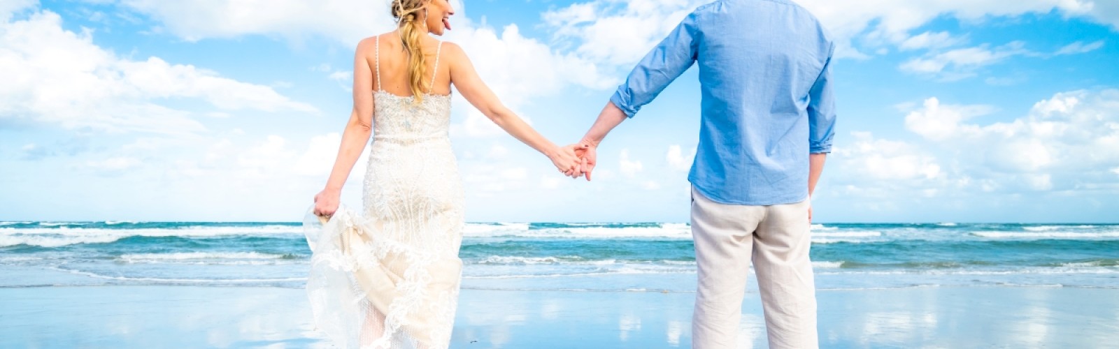 A couple holds hands while standing on a sandy beach, facing the ocean, with reflections on the wet sand and a bright blue sky above.