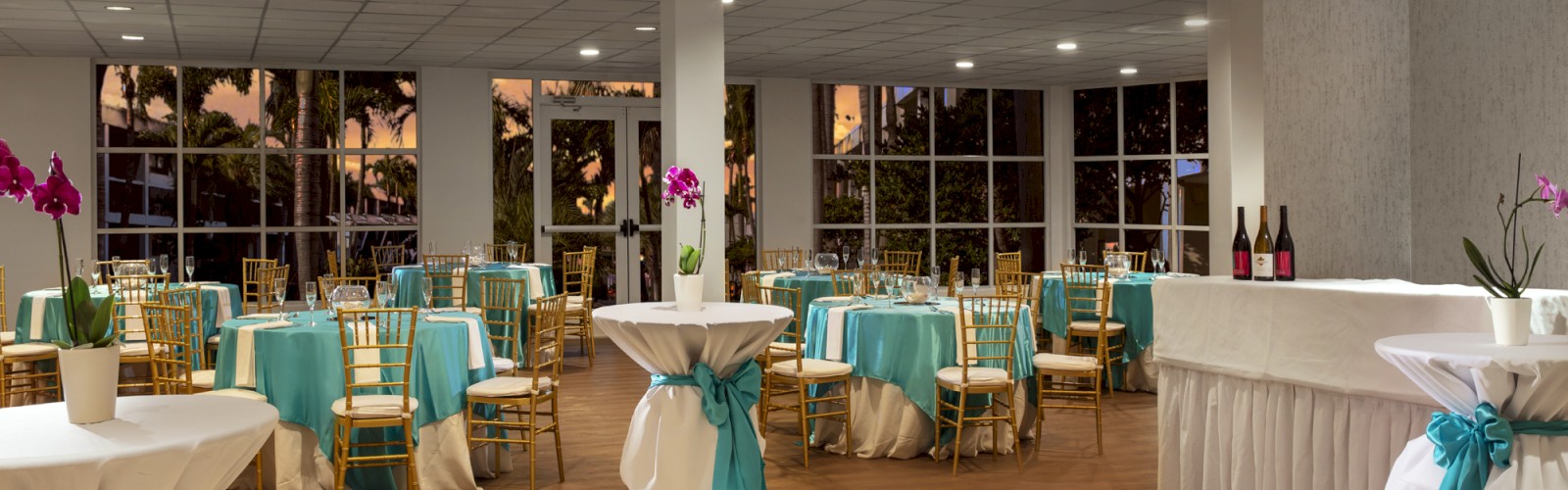 A decorated event hall with tables and chairs covered in teal and white cloths, high cocktail tables, and bottles on a counter.