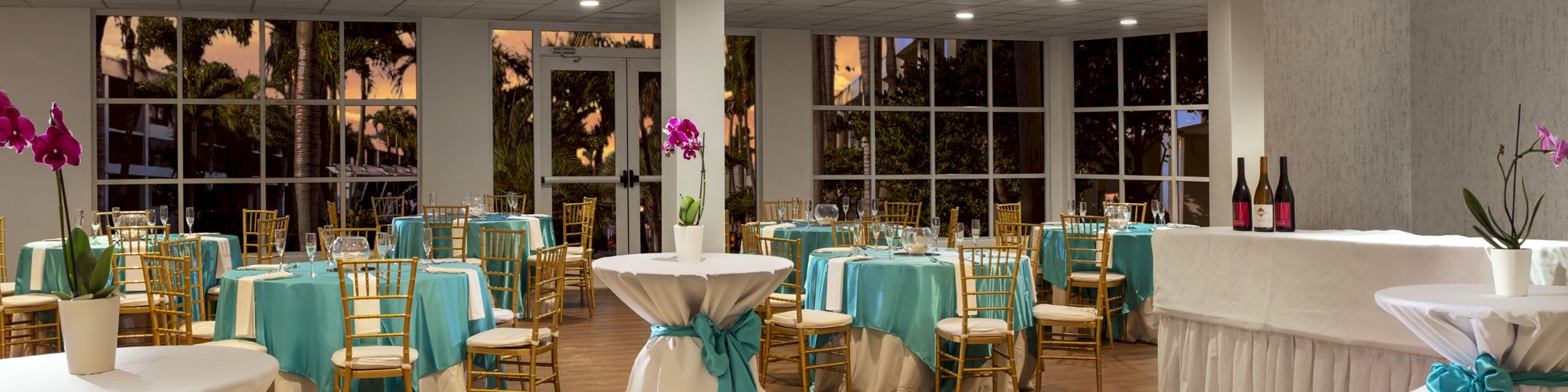 A decorated event hall with tables and chairs covered in teal and white cloths, high cocktail tables, and bottles on a counter.