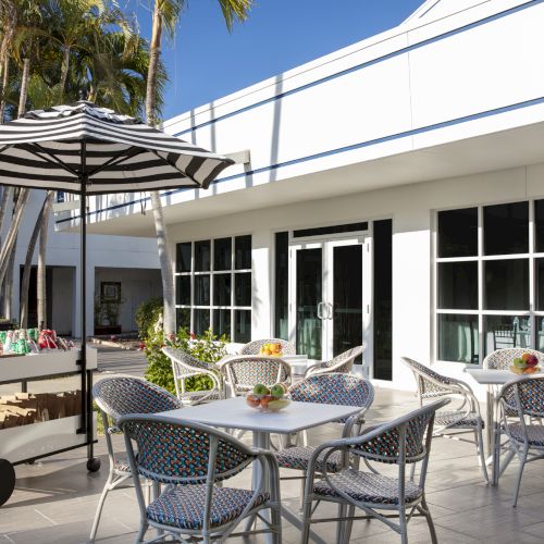 An outdoor patio features wicker chairs, small round tables with drinks, a cart with an umbrella, and a modern white building in the background.