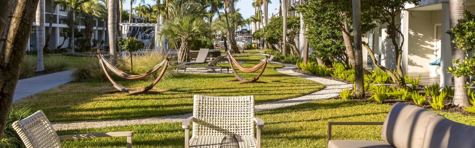 A landscaped outdoor area at a resort features chairs, a table, palm trees, and hammocks strung between trees on a sunny day.