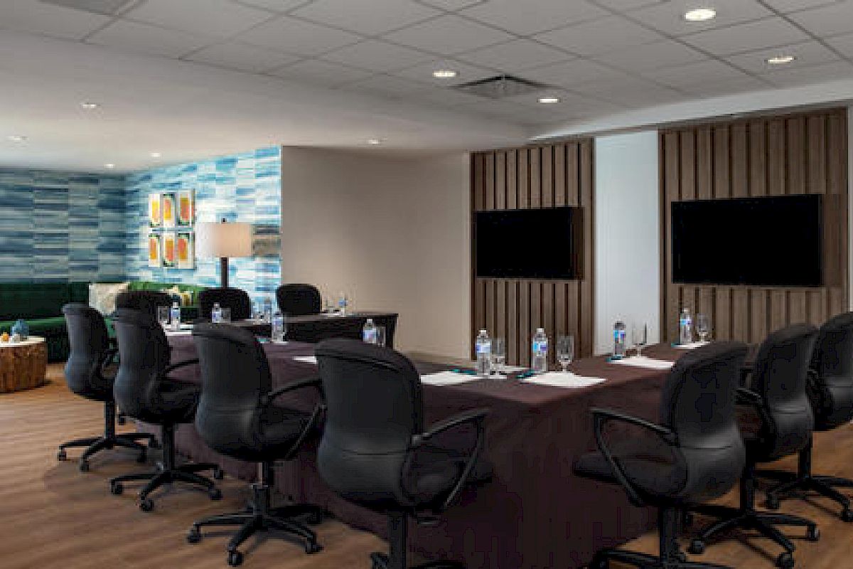 The image shows a conference room with a table and twelve chairs, water bottles, notepads, and two screens on the wall.