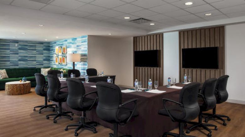 The image shows a conference room with a table and twelve chairs, water bottles, notepads, and two screens on the wall.