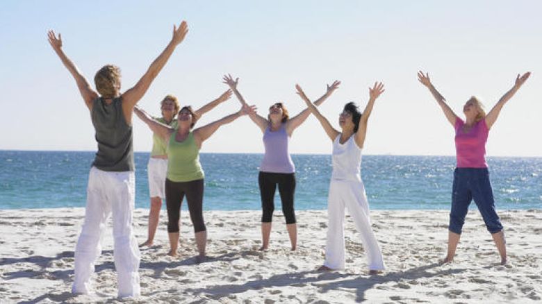 A group of people are on a beach, stretching with their arms raised, facing an instructor against a backdrop of the ocean and blue sky.