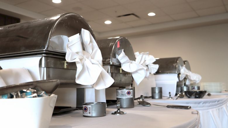 Chafing dishes with white cloth decorations on a buffet table, along with various condiments and utensils, are seen in the image.