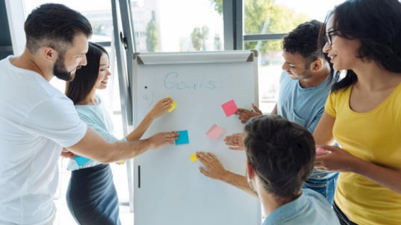 A diverse group of people are collaborating, placing sticky notes on a whiteboard during a brainstorming session in a well-lit room.