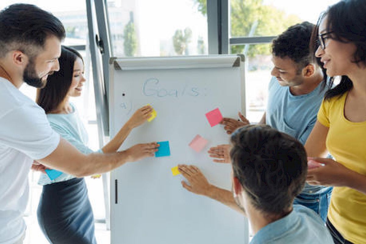 A diverse group of people are collaborating, placing sticky notes on a whiteboard during a brainstorming session in a well-lit room.