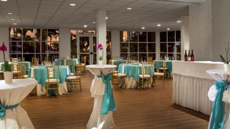 A decorated event room with round tables covered in teal and white, high-top tables, a bar area with wine bottles, and large windows in the background.