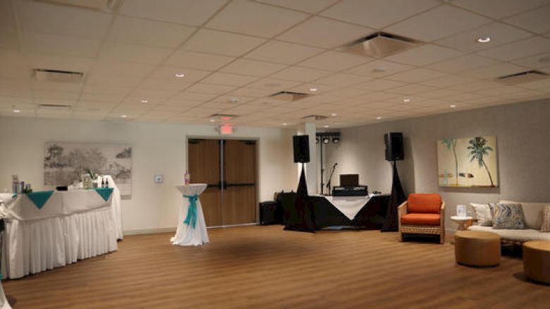 The image shows an empty, decorated room with tables, a DJ setup, and seating areas, likely set up for an event or party.