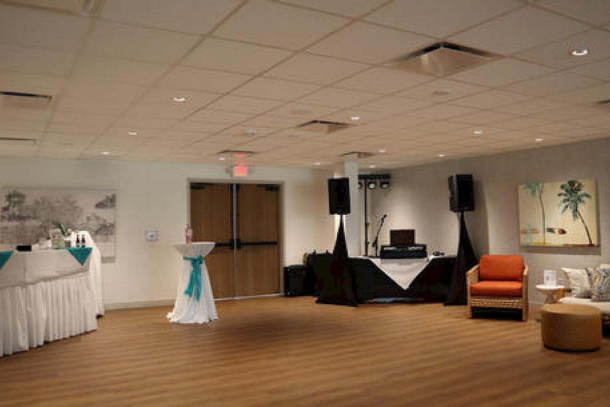 The image shows an empty, decorated room with tables, a DJ setup, and seating areas, likely set up for an event or party.