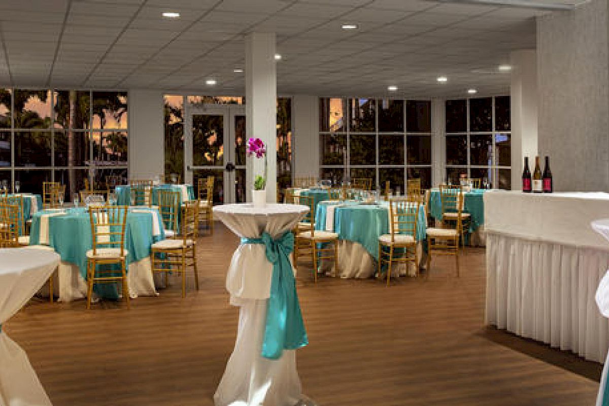 The image shows an elegant event space with round tables covered in turquoise and white tablecloths, gold chairs, and standing tables, all set for a function.