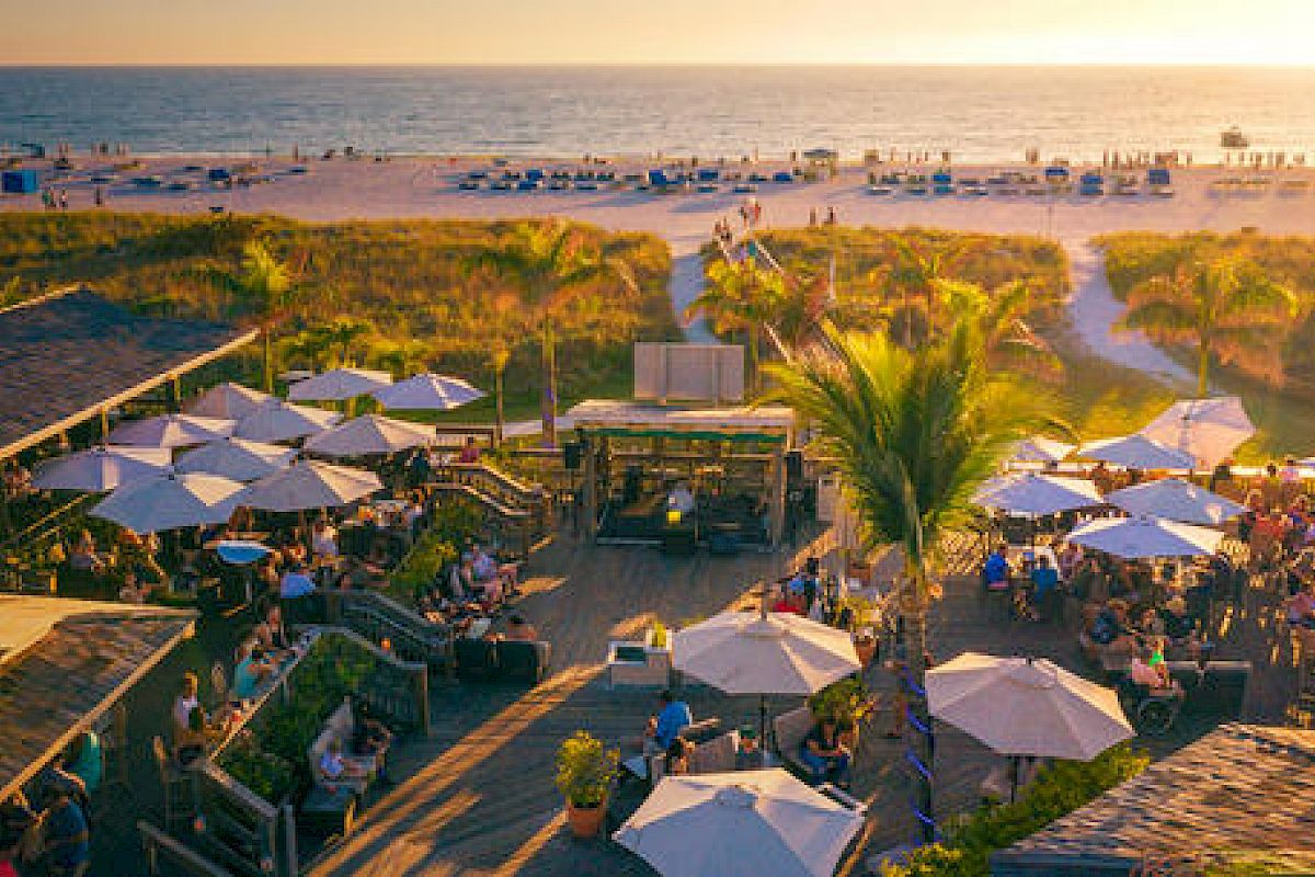 A lively beachfront scene with people gathered under umbrellas, a wooden deck, a pathway to the beach, soft lighting from a setting sun.