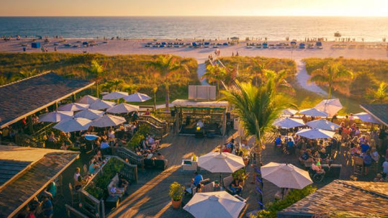 A lively beachfront scene with people gathered under umbrellas, a wooden deck, a pathway to the beach, soft lighting from a setting sun.