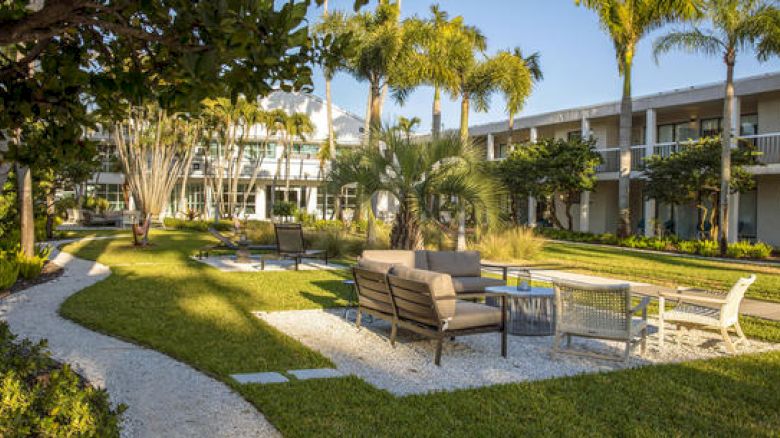 The image shows an outdoor courtyard with seating arrangements, palm trees, and a path winding through. In the background, there is a two-story building.