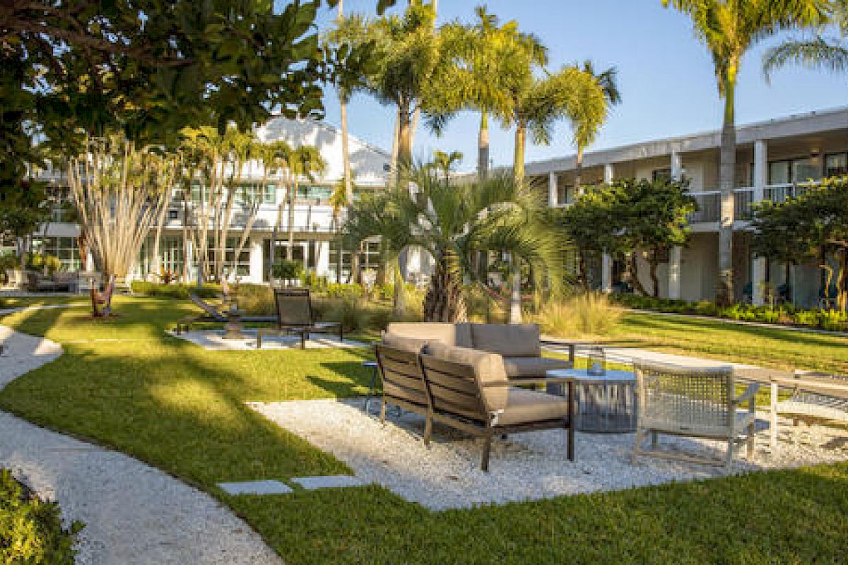 The image shows an outdoor courtyard with seating arrangements, palm trees, and a path winding through. In the background, there is a two-story building.