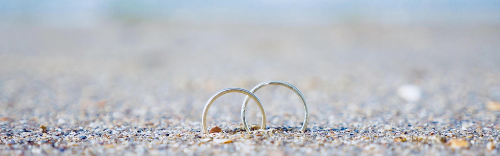 Two silver rings placed on a sandy beach with a blurred background of water, symbolizing love or commitment.