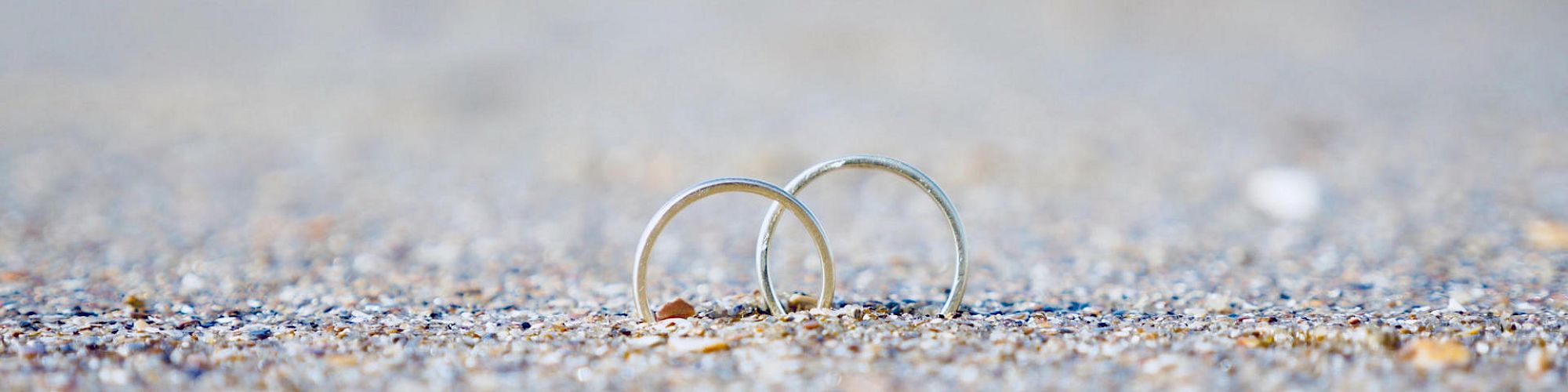 Two silver rings placed on a sandy beach with a blurred background of water, symbolizing love or commitment.