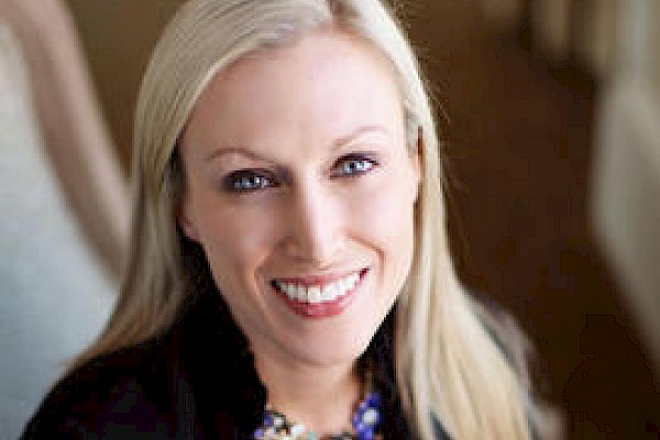 A smiling woman with blonde hair wears a black jacket and a colorful necklace, sitting in what appears to be an indoor setting.