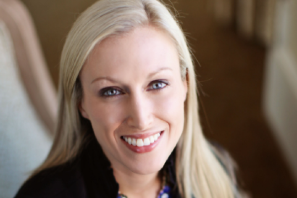 A person with long blonde hair, wearing a black blazer and colorful necklace, is smiling while seated indoors.