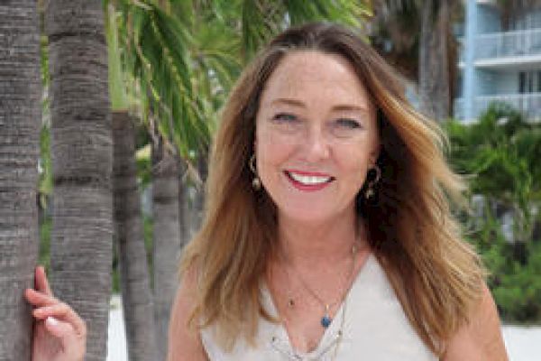 A woman stands by palm trees, smiling, wearing a sleeveless beige dress, accessorized with jewelry. There is greenery and a building in the background.