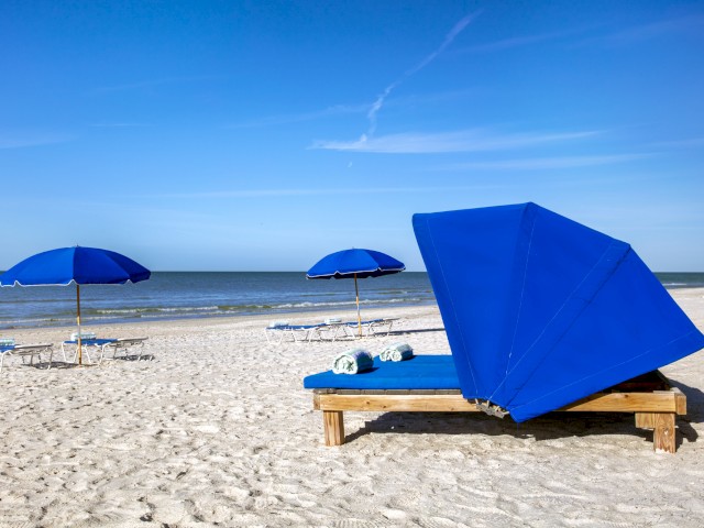 A tranquil beach scene with sunbeds, blue umbrellas, clear sky, and calm sea.