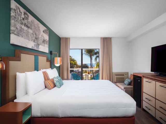 A neat hotel room with a comfy bed, dresser, TV, and a view of palm trees outside.