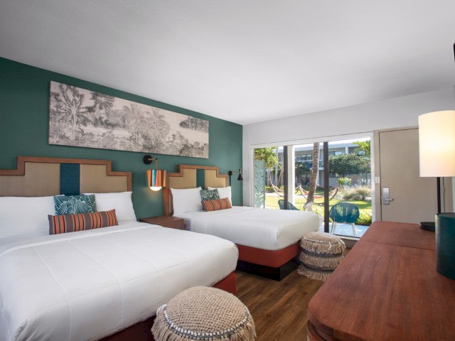 A modern hotel room with two beds, nightstand, lamp, and a desk with a view outside.
