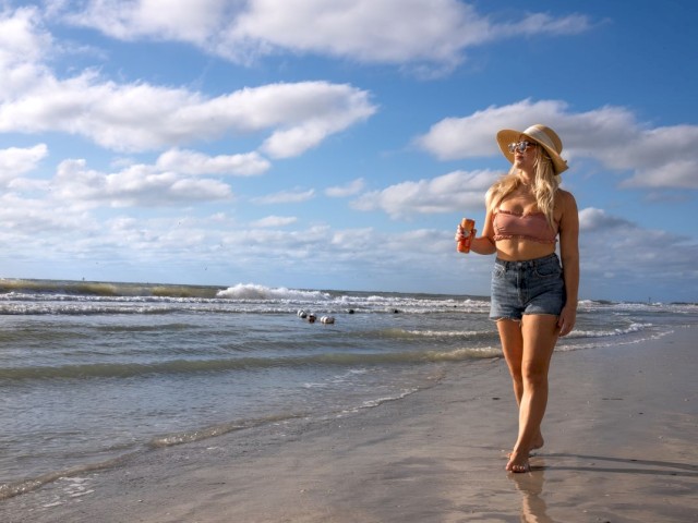 A person is walking on a sunny beach with a drink, wearing a hat and sunglasses.