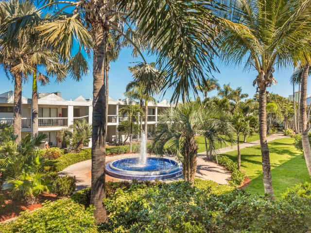 A tropical resort setting with palm trees, a fountain, and two-story buildings.