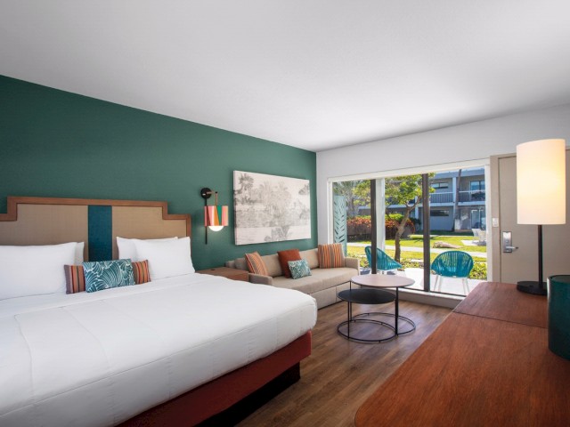 Modern hotel room with a bed, seating area, artwork on the wall, and view of the outdoors.