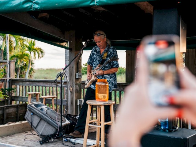 A person is taking a photo of a guitarist performing outdoors.