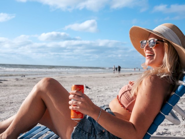 A person is relaxing on a beach chair by the sea, holding a drink, and wearing a hat and sunglasses.