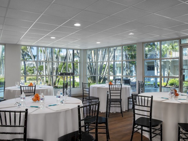 Bright dining room with tables set for a meal, surrounded by large windows showing lush greenery.
